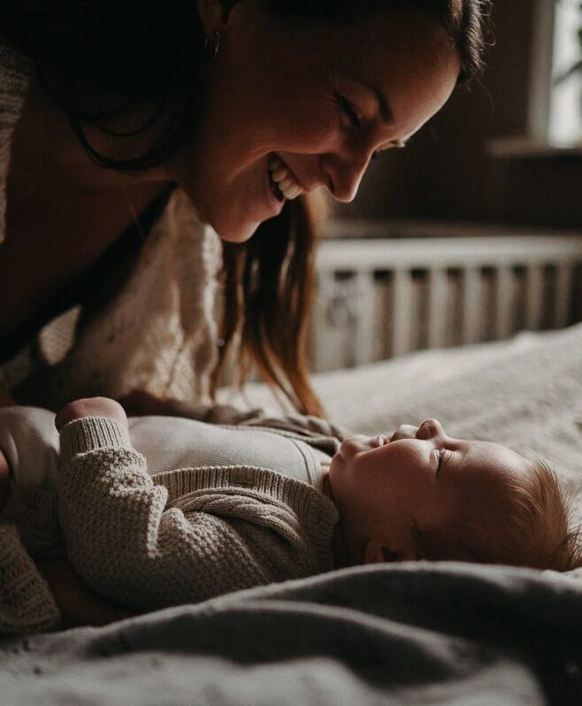 Bebis ligger på sängen och mamman lutar sig fram emot hen och ler.